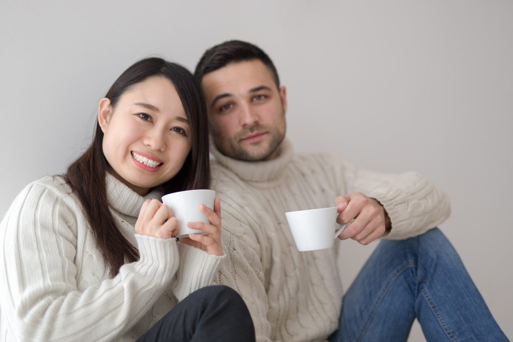 An Asian woman and white male partner sit together drinking coffee, She rests her head on his shoulder.